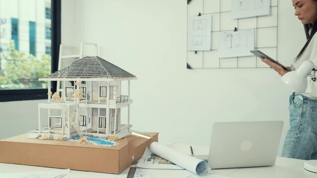 Beautiful caucasian architect focus on using tablet to check house model on meeting table with blueprint, house model and architectural document scatter around at meeting room. Immaculate.