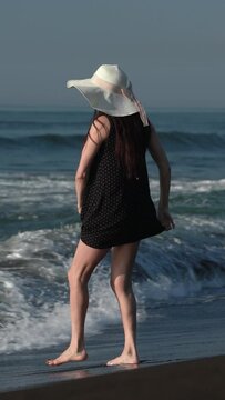 Barefoot mature adult woman walking on black sandy beach on Pacific Ocean. Full length Caucasian ethnicity female dressed in black summer polka dot dress and straw sun hat. Vertical video, handheld