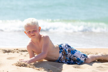 A smiling little boy lies on a beach against the backdrop of the sea and blue sky. Little boy lying on the sand and playing on the beach of Mediterranean sea. Summertime, vacation, travel banner.