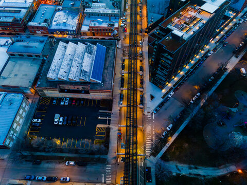 aerial drone view of Urban streets, bathed in warm twilight hues, form a symmetrical pattern. Buildings with textured rooftops flank the illuminated roads. A tranquil view from above, 