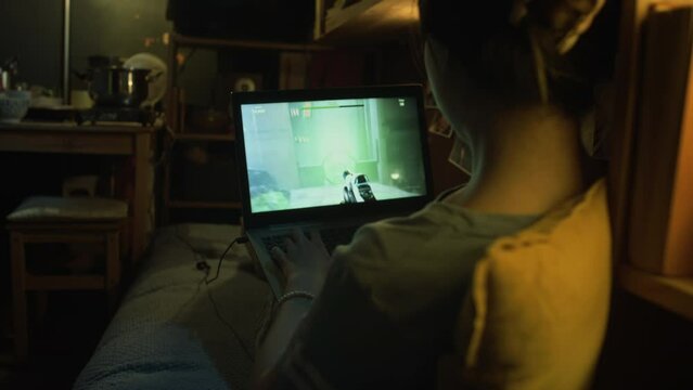 Medium rear footage of anonymous young female sitting on bed in cluttered shoebox flat in evening, playing shooter video game on laptop computer online, passing level