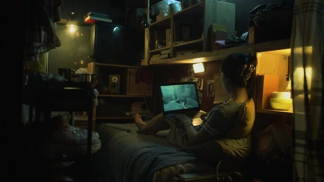 Wide rear footage of unrecognizable young woman sitting on bed in shabby micro flat at nighttime, playing shooter video game on laptop computer, steaming pot on stove under light bulb in corner