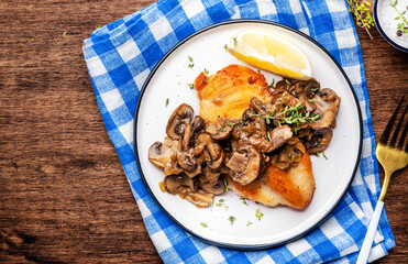 French breaded chicken steak with mushrooms and onion sauce with white wine, lemon and thyme on plate, rustic table background, top view