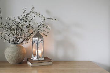 White flowers, blooming prunus tree branches in vase. Glowing Moroccan lantern on books. Wooden...