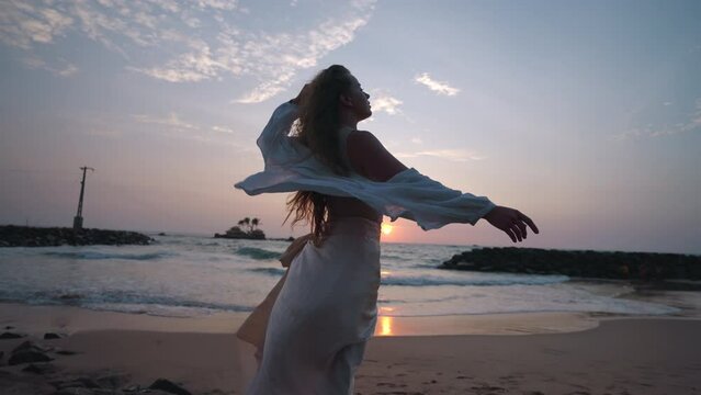 Pretty curly girl posing, long hair blowing in wind at amazing sunset on beach. Young woman enjoying summer sunrise at seaside in morning, windy weather. Caucasian model catches a wind at ocean shore.