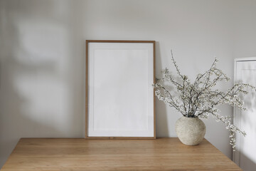 Vertical wooden picture frame, poster mockup in sunlight. Spring composition. Elegant interior, home office still life. Artistic display. Blooming cherry plum tree branches in vase. Wooden table, desk