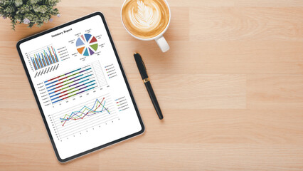 A digital tablet on a wooden desk displaying colorful business analytics graphs alongside a cup of coffee and a pen.