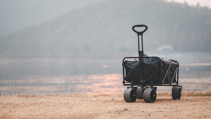 A black utility cart stands alone on a sandy lakeshore, against a backdrop of soft twilight hues. - obrazy, fototapety, plakaty