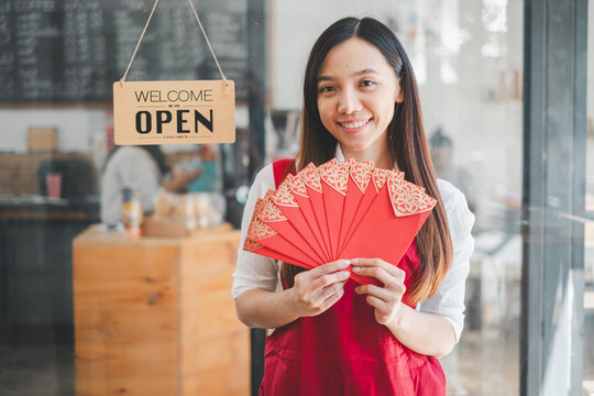 Happy Asian woman entrepreneur holding red envelopes, ready to celebrate, standing in front of a welcoming open sign at her cafe.