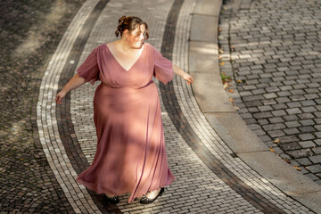 A young plus size woman wearing a pink dress dancing in the street of a city. Body positivity...