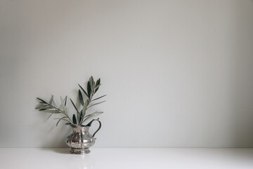 Olive tree branches, twigs in silver jug vase on white table. Empty mint green wall mockup, background. Working space, home office decor. Vintage Mediterranean summer interior still life.