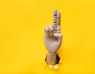 A wooden hand protrudes from a torn hole in yellow paper and shows two fingers. Concept of...
