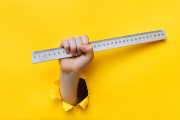 A child's hand reaches through a torn hole in yellow paper and holds a wooden ruler. The concept of teaching geometry and mathematics at an early age.