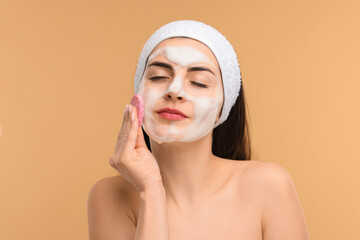 Young woman with headband washing her face using sponge on beige background