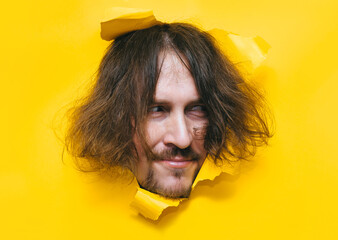 A long-haired, shaggy man with a beard and mustache looks out from a torn hole in yellow paper with...