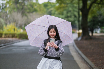Obraz premium 雨の日に傘をさしてスマートフォンを見ながら歩く小学生の女の子