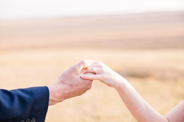 An engaged couple (man and woman) are in a dry field holding hands. 