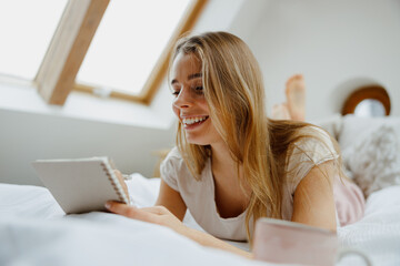 Blond happy woman making notes in note pad while sitting on bed at home 