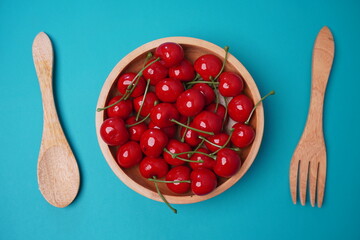 cherries in a wooden plate between spoon and fork on isolated blue background table
