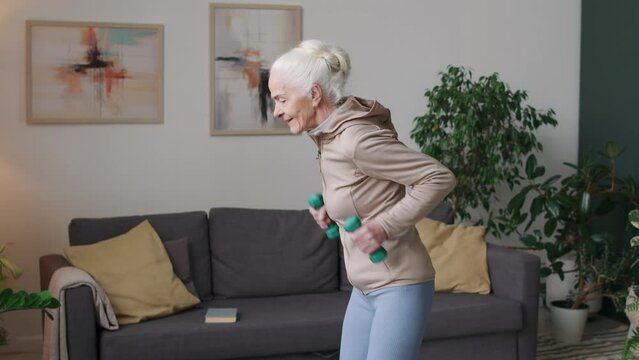 Medium shot of Caucasian senior woman doing triceps kickback exercise with dumbbells while having indoor fitness training in apartment at daytime