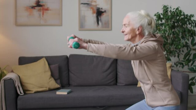 Tilt up shot of cheerful Caucasian elderly woman squatting down with two dumbbells in hands during indoor training in modern apartment