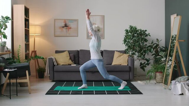 Full stab shot of Caucasian elderly woman practicing yoga standing in asana with praying hands above her head on mat in modern apartment