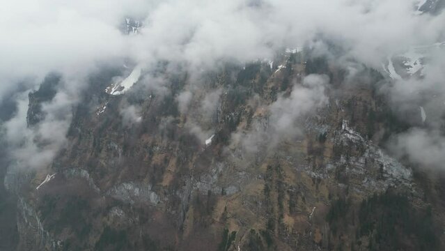 Panoramic overview of wispy tendrils of clouds up against sheer mountain cliffs covered in snow