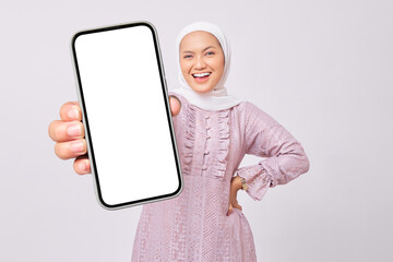 Smiling beautiful young Asian Muslim woman wearing hijab and purple dress showing mobile phone with...