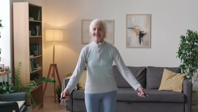 Tilting up shot of cheerful Caucasian aged woman jumping with rope while having cardio training at home in morning