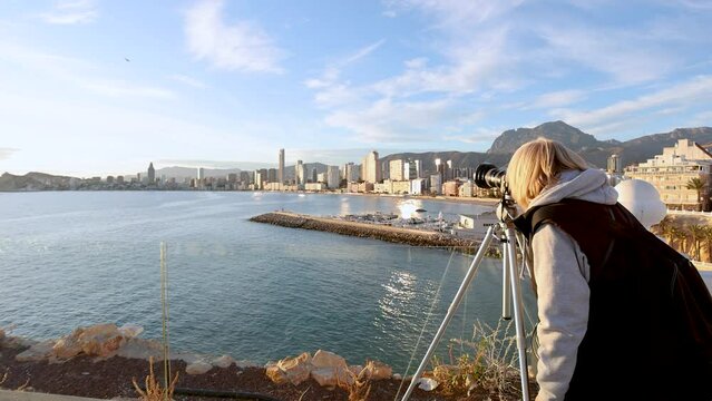 Blonde woman photographer taking photos of Benidorm marina and skyline using a tripod