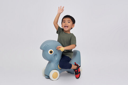 Full body little small smiling happy Asian boy 2-3 years old wearing green t-shirt  sitting on toy rocking horse and waving hand on friend isolated on white background