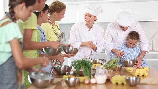 Amiable woman and young man, skilled chefs wearing white cook jackets and toques, running culinary courses for preteen children, sharing secrets of cooking 