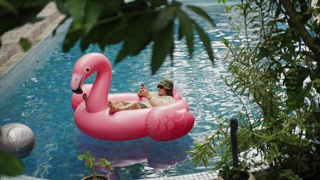 Man use phone on inflatable pink flamingo float mattress on pool party. 