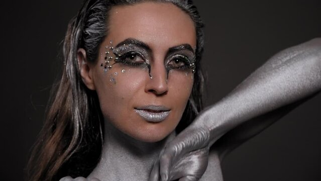Close-up of a female model with silver skin and hands, with bright makeup with stones posing in the studio on a black background.