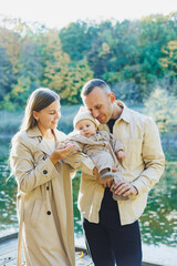 Fototapeta premium A happy young couple with a little son in their arms in the park near the lake. Family vacation in nature with a small son. The concept of family vacation.