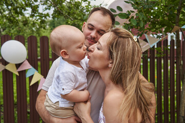 Fototapeta premium Mom kisses the baby, who is sitting in Dad's arms. happy young family surrounded by green trees and festive flags