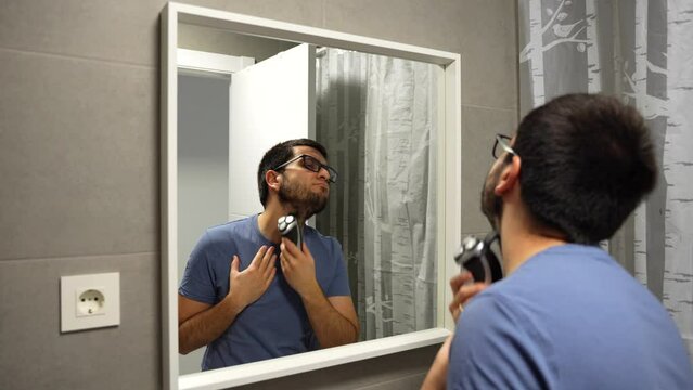 Man with glasses shaving his beard with machine in bathroom mirror