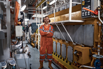 Young, smiling engineer in orange boiler suit, standing near main engine in engine room.