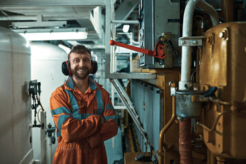 Smiling engineer in orange boiler suit, standing near main engine control box in engine room.