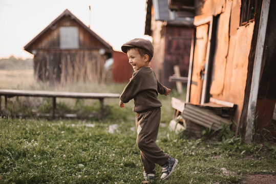 A boy is playing on a farm or ranch field, resting. Portrait of a little boy in a cap and overalls. Childhood. An authentic picture with candid emotions.