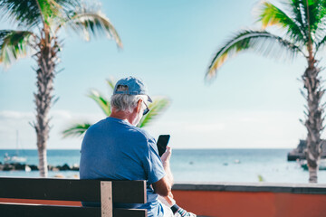 Back view of senior man using smartphone sitting outdoors on bench face the sea enjoying sunny day, travel, vacation, retirement lifestyle
