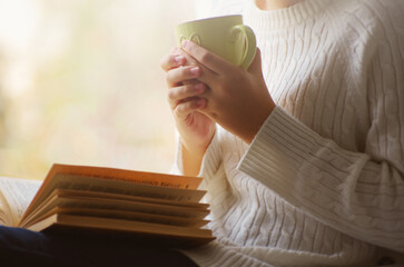 Girl is holding cup of tea and reading a book beside the window