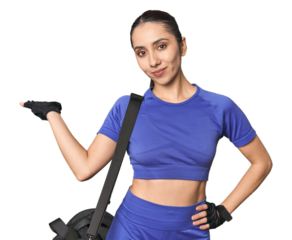 Athletic Caucasian young woman with gym bag on studio background showing a copy space on a palm and holding another hand on waist.