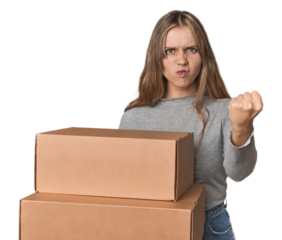 Blonde woman with cardboard boxes moving on studio backdrop showing fist to camera, aggressive facial expression.