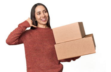 Caucasian young woman with moving boxes on studio background showing a mobile phone call gesture with fingers.