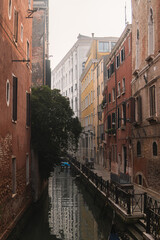 Tranquil Venetian canal scene with charming brick and colorful buildings, a lush tree leans over the calm water, where the scene is reflected, with a boat gently moored by the quay, on a cloudy day.