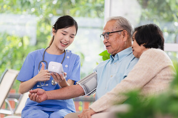 A smiling nurse takes a senior man's blood pressure, an elderly woman beside him, in a bright, plant-filled room. Healthcare compassion and elderly patient care concept.
