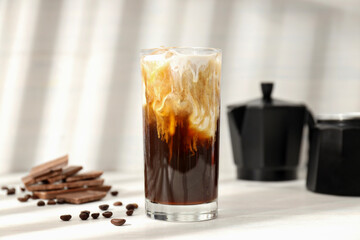 Glass with iced coffee, coffee maker and coffee bean on light background, close up