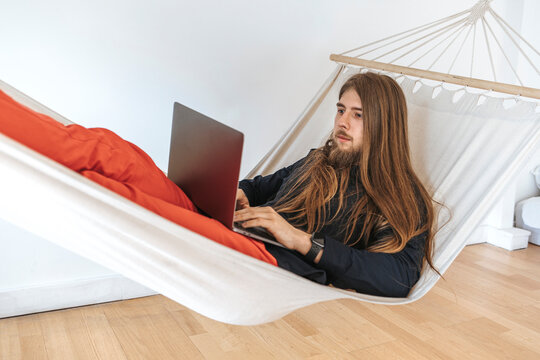 Young programmer on hammock using laptop for coding at startup in office
