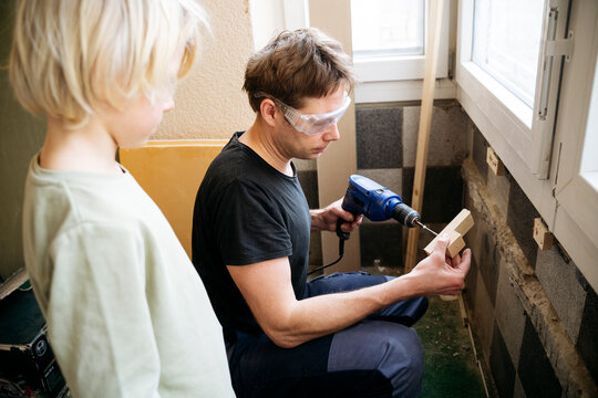 Mature man drilling wooden piece near son at home